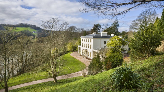 View through a wintry garden with bare trees and the River Dart peeping between them, to the white façade of Greenway House